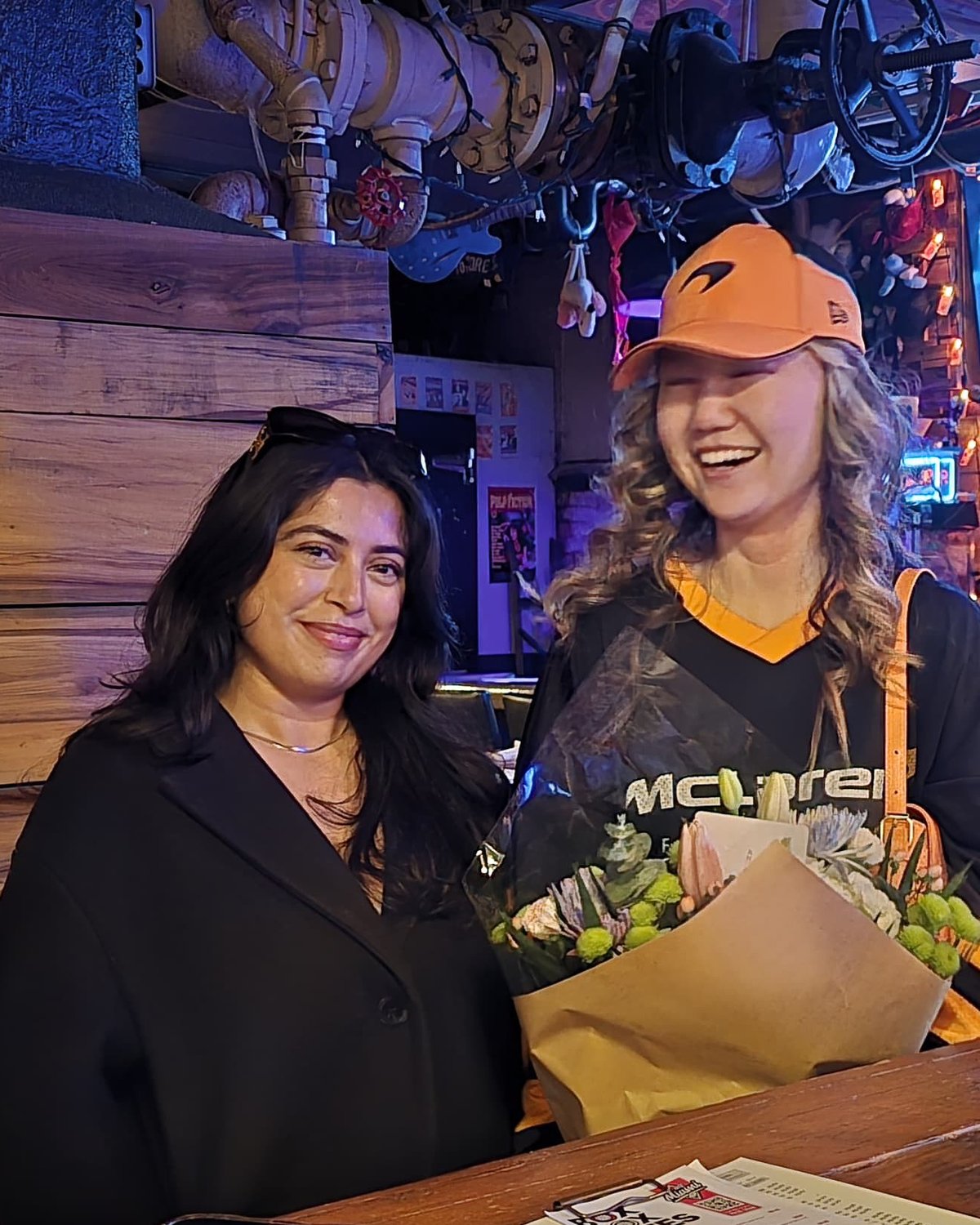 Two friends at the bar on race day, one in her McLaren cap and shirt with a bunch of flowers